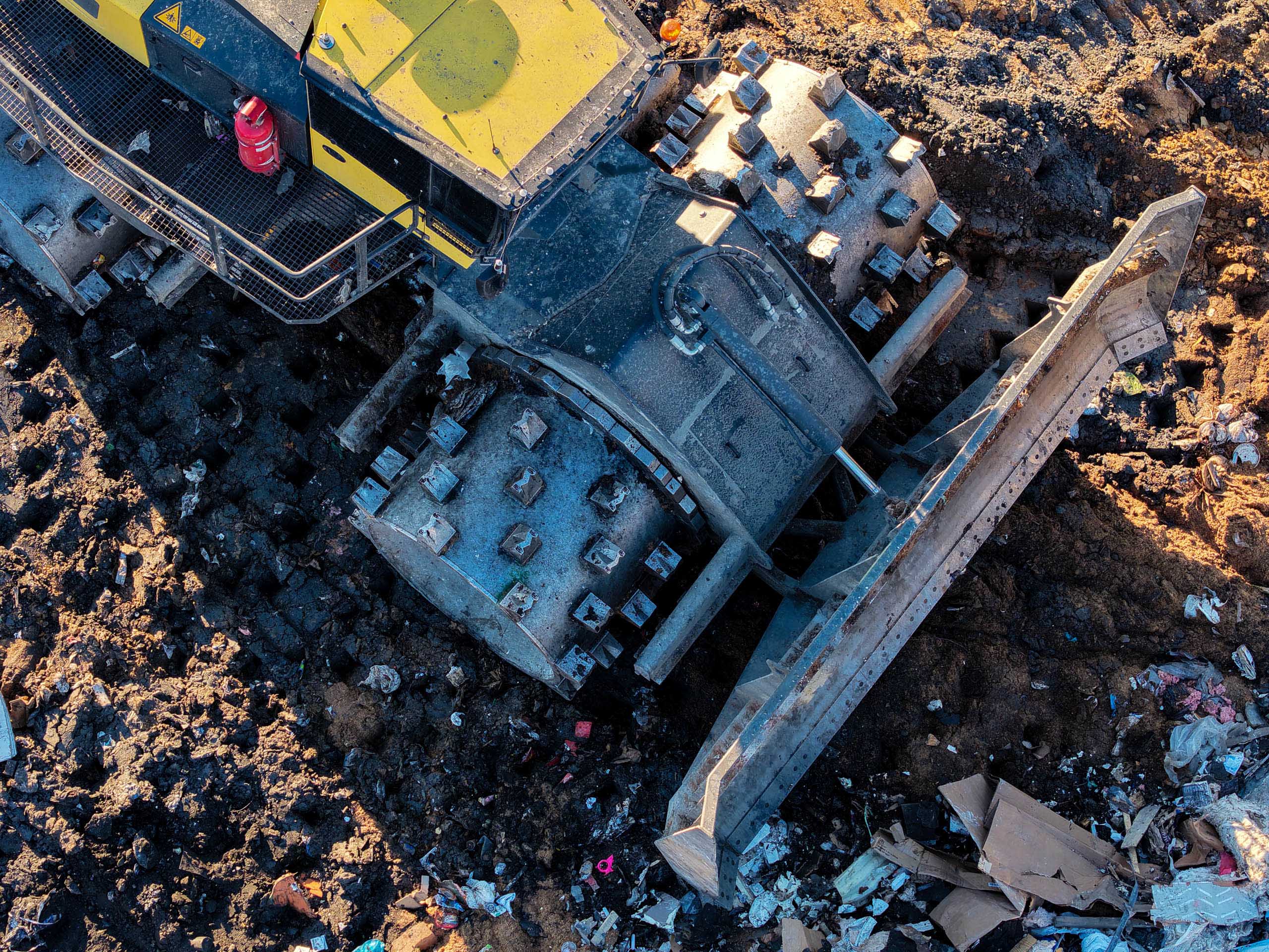 Aerial view of a yellow bulldozer with a worker in red standing on it, pushing debris and garbage on the muddy Frey Farm Landfill site, highlighting waste management efforts.
