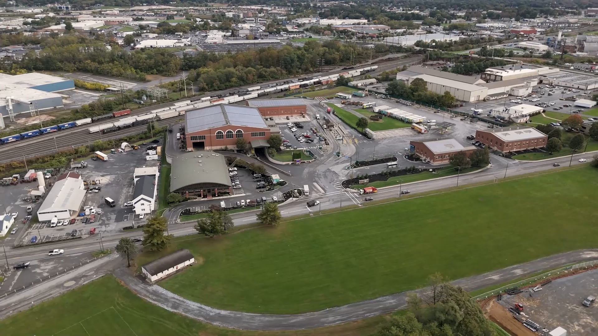 Aerial view of a lcswma area with large green fields, several buildings, parking lots, roads, and a train passing by on tracks in the background. Trees and a Waste Management transfer station are visible near the industrial areas further out.