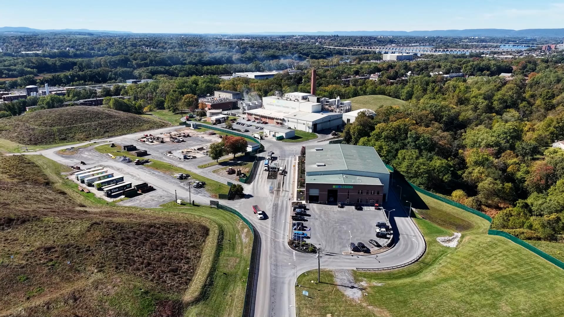 Aerial view of the Lancaster Waste-To-Energy facility surrounded by green fields and trees, with several buildings, parking lots, trucks, and roads, set against a backdrop of forested hills and a distant cityscape.