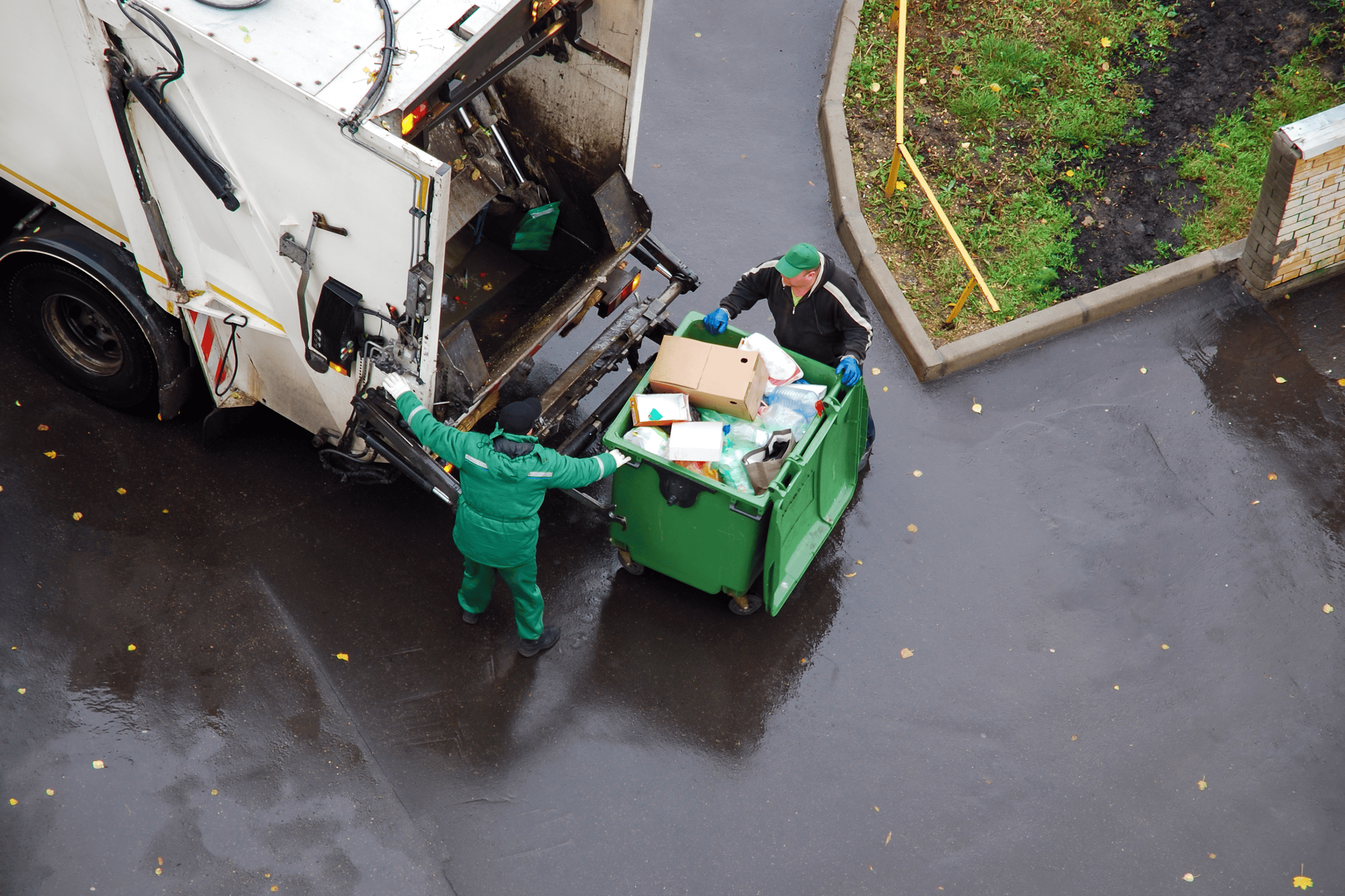 Two trash workers in green uniforms empty a large green garbage bin into a white garbage truck on a wet street near a grassy area.