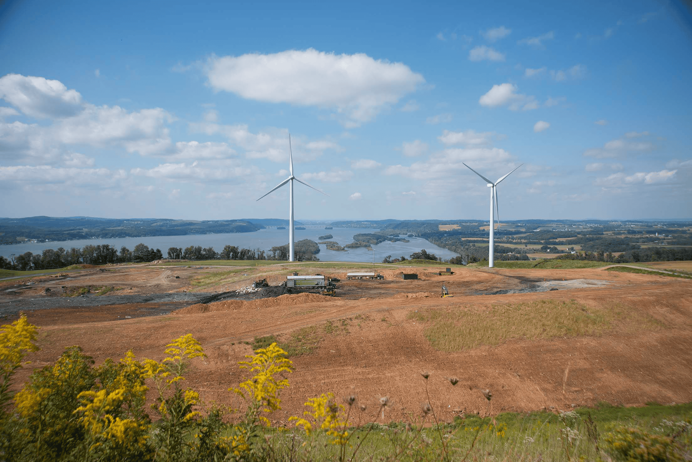 Two large wind turbines stand on a grassy hill overlooking a river and distant hills, with a partly cloudy sky above. Construction vehicles and cleared land are visible in the foreground.