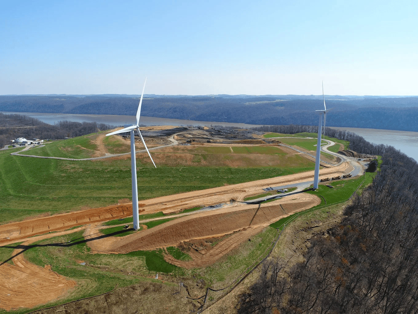 Aerial view of two large wind turbines on a grassy hill near a river, with winding roads, scattered trees, and distant rolling hills under a clear blue sky.