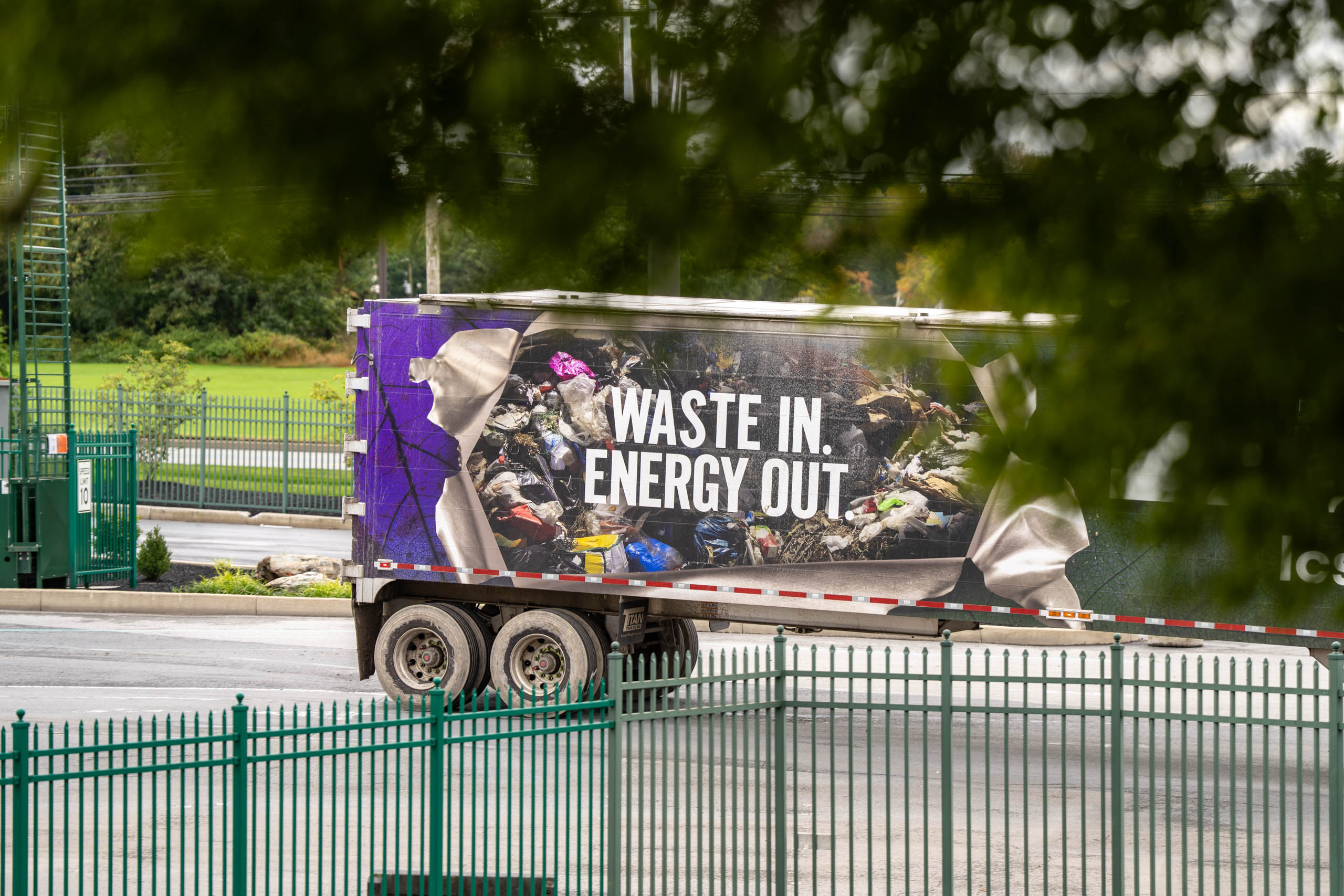 A lcswma truck with an image of piled trash and the words WASTE IN. ENERGY OUT is parked near a green fence, partially framed by tree branches.