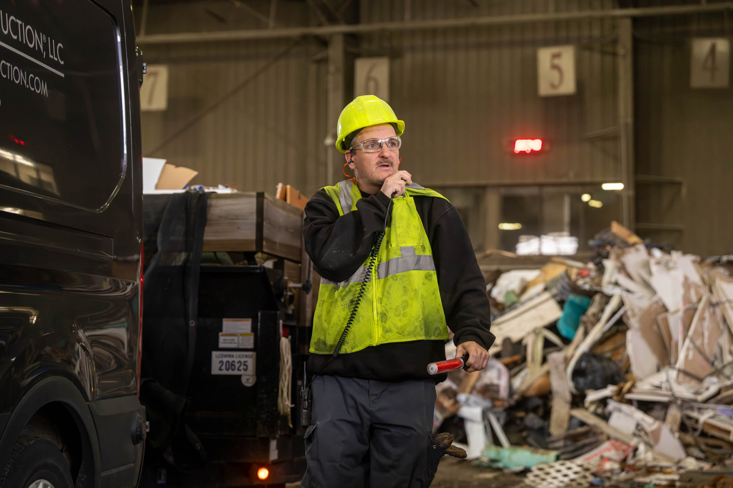 A worker wearing a yellow hard hat and reflective vest talks into a walkie-talkie in a large lcswma facility, surrounded by piles of debris and construction waste, likely monitoring Residential Disposal Rates during site operations.