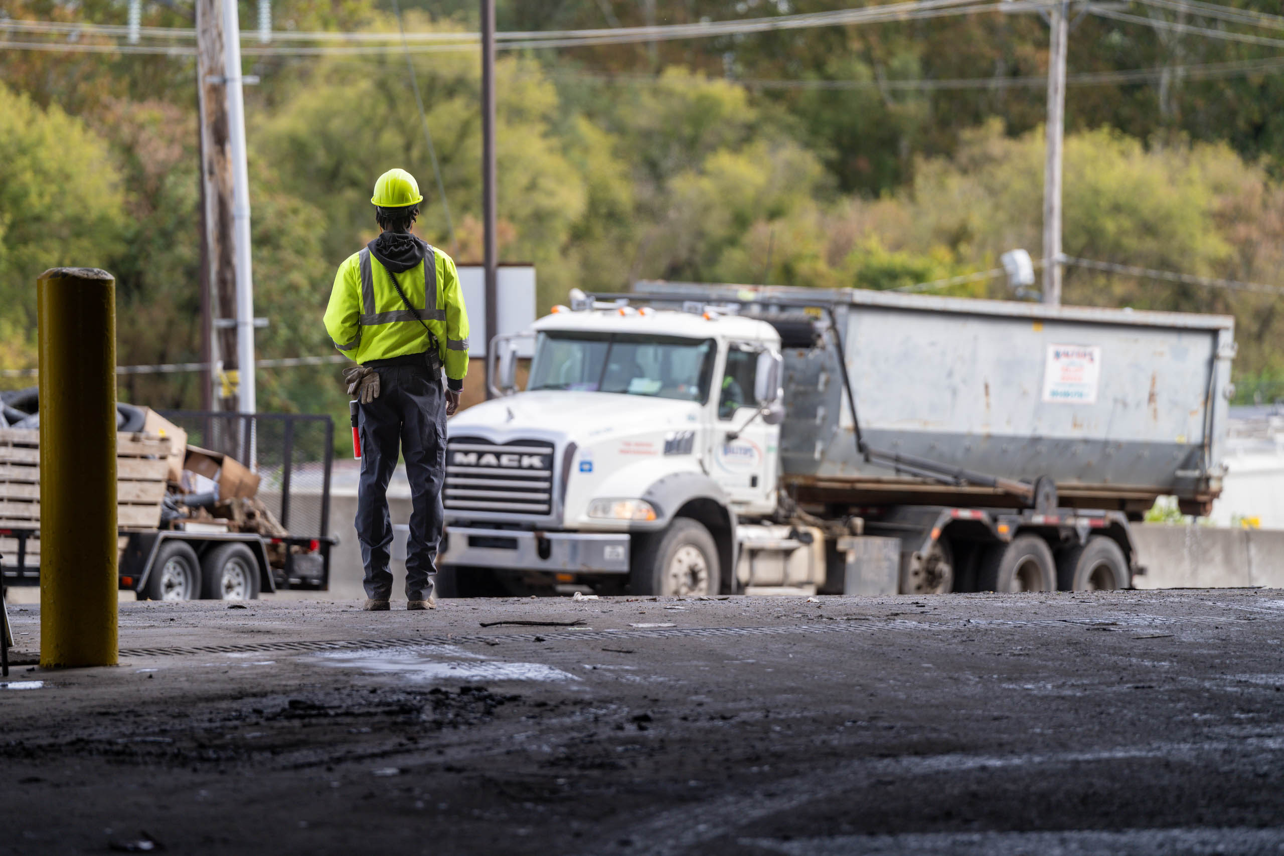 A lcswma worker in a yellow safety vest and helmet stands facing a white dump truck on a worksite, where Residential Disposal Rates influence the project's efficiency, with trees and power lines seen in the background.