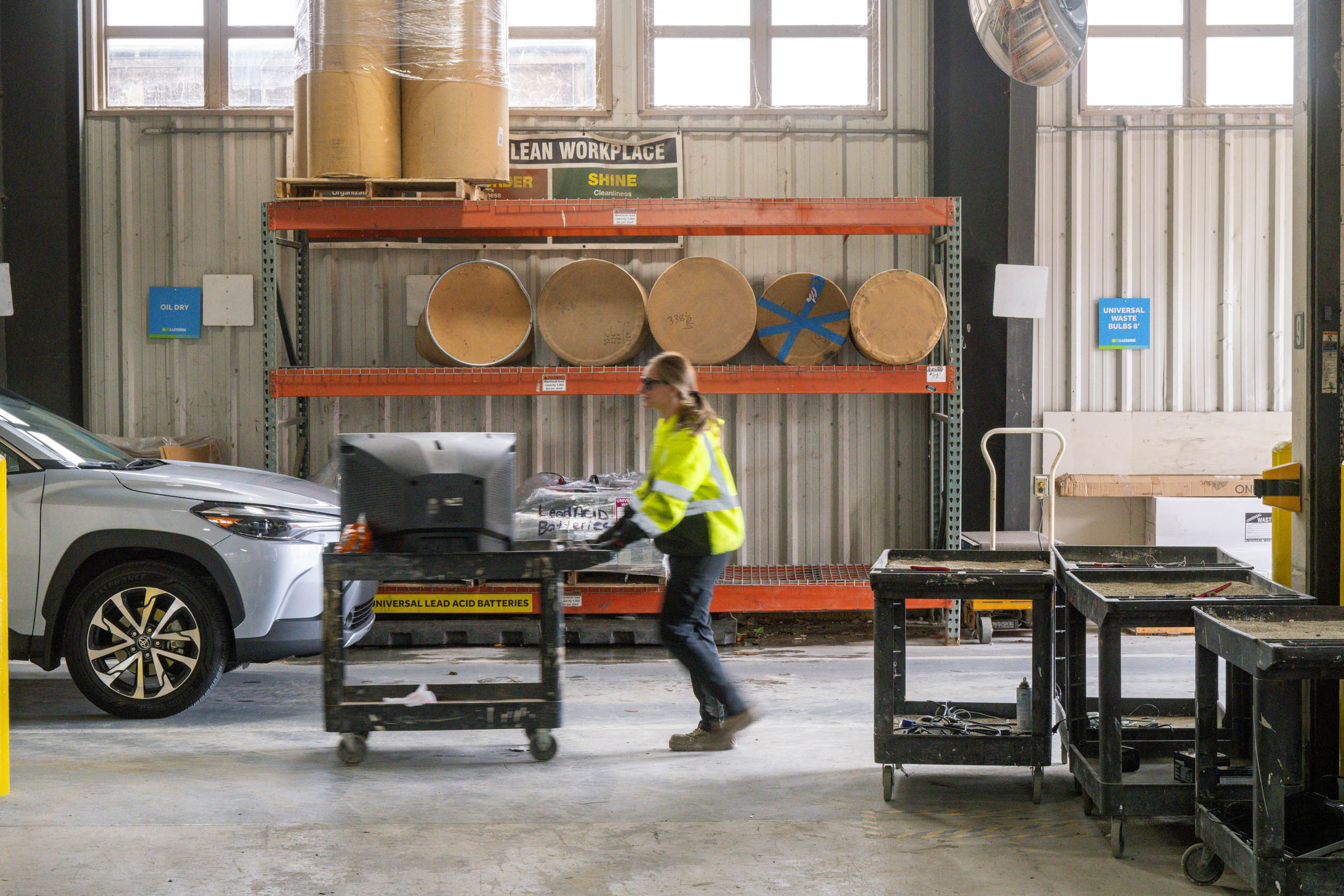 lcswma person in a yellow safety jacket pushes a cart with electronic equipment through a warehouse area, surrounded by industrial shelves, large drums, and a parked car—highlighting logistics behind managing residential disposal rates.