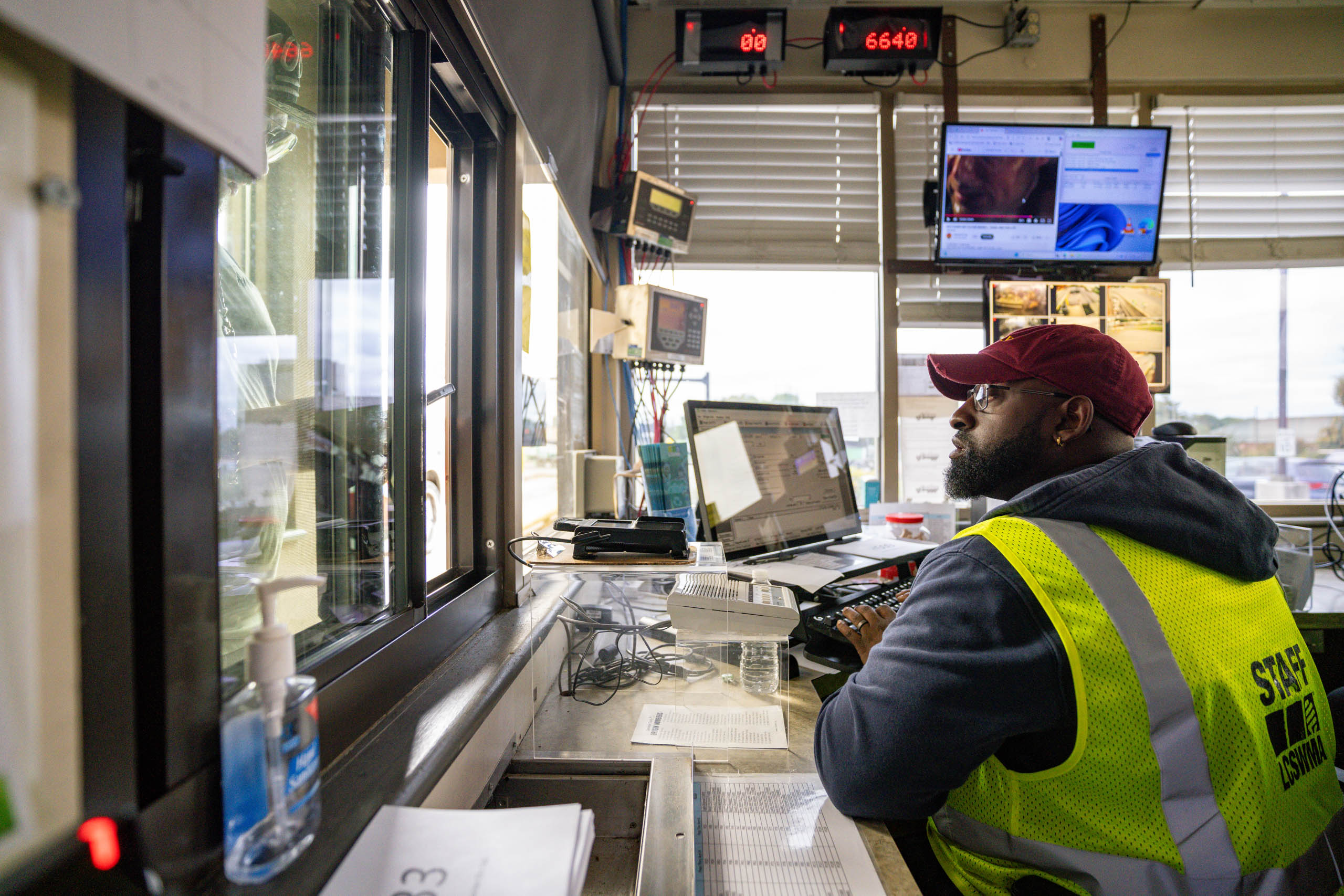 A man in a yellow safety vest and red cap sits at a desk by a window in lcswma office filled with monitors, computers, and technical equipment, attentively monitoring residential disposal rates as he looks outside.