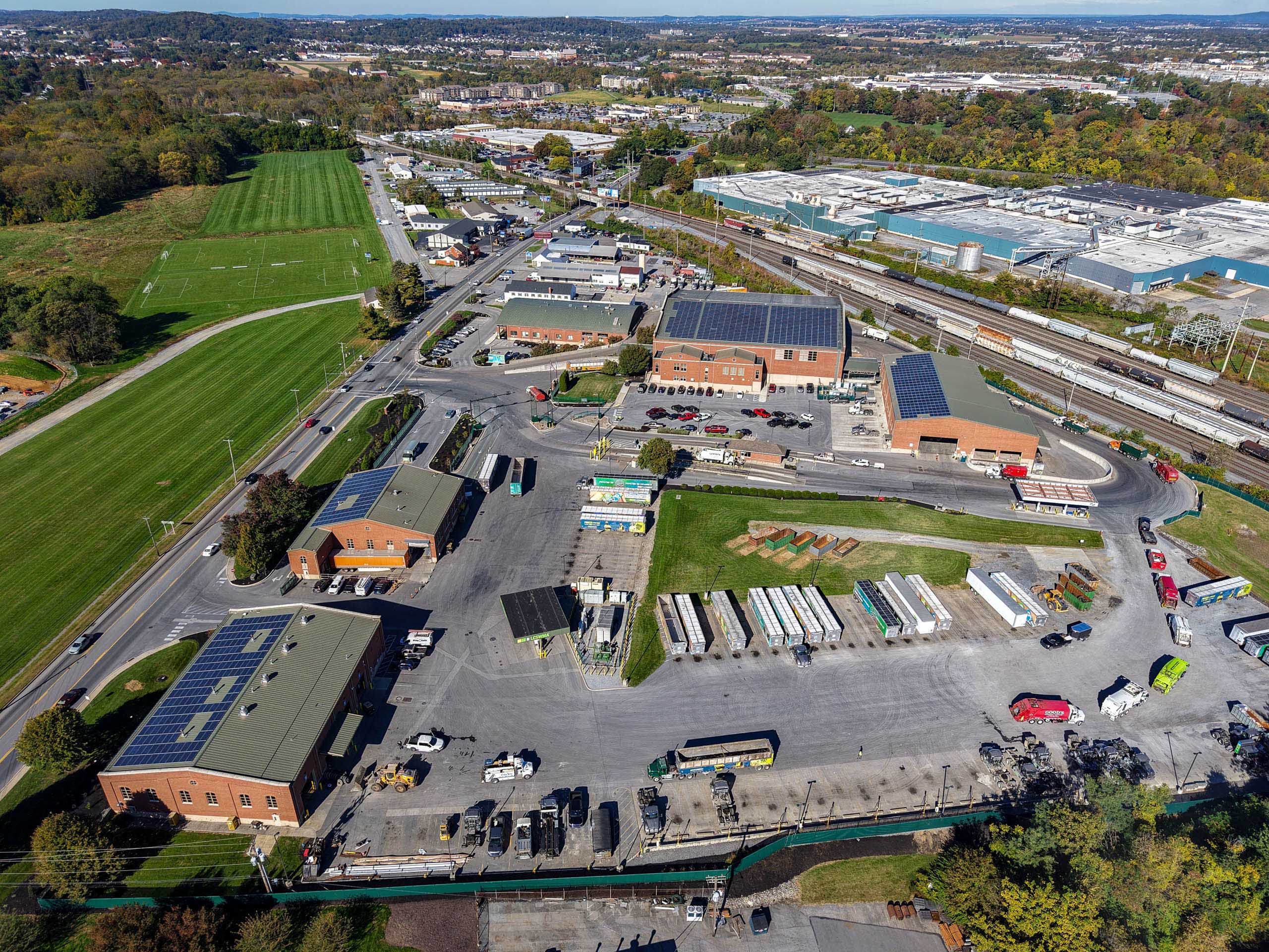 Aerial view of an lcswma park with warehouses, parking lots, trucks, and buildings with solar panels. Green sports fields and trees are visible on the left, with roads and a rail line running through the area.