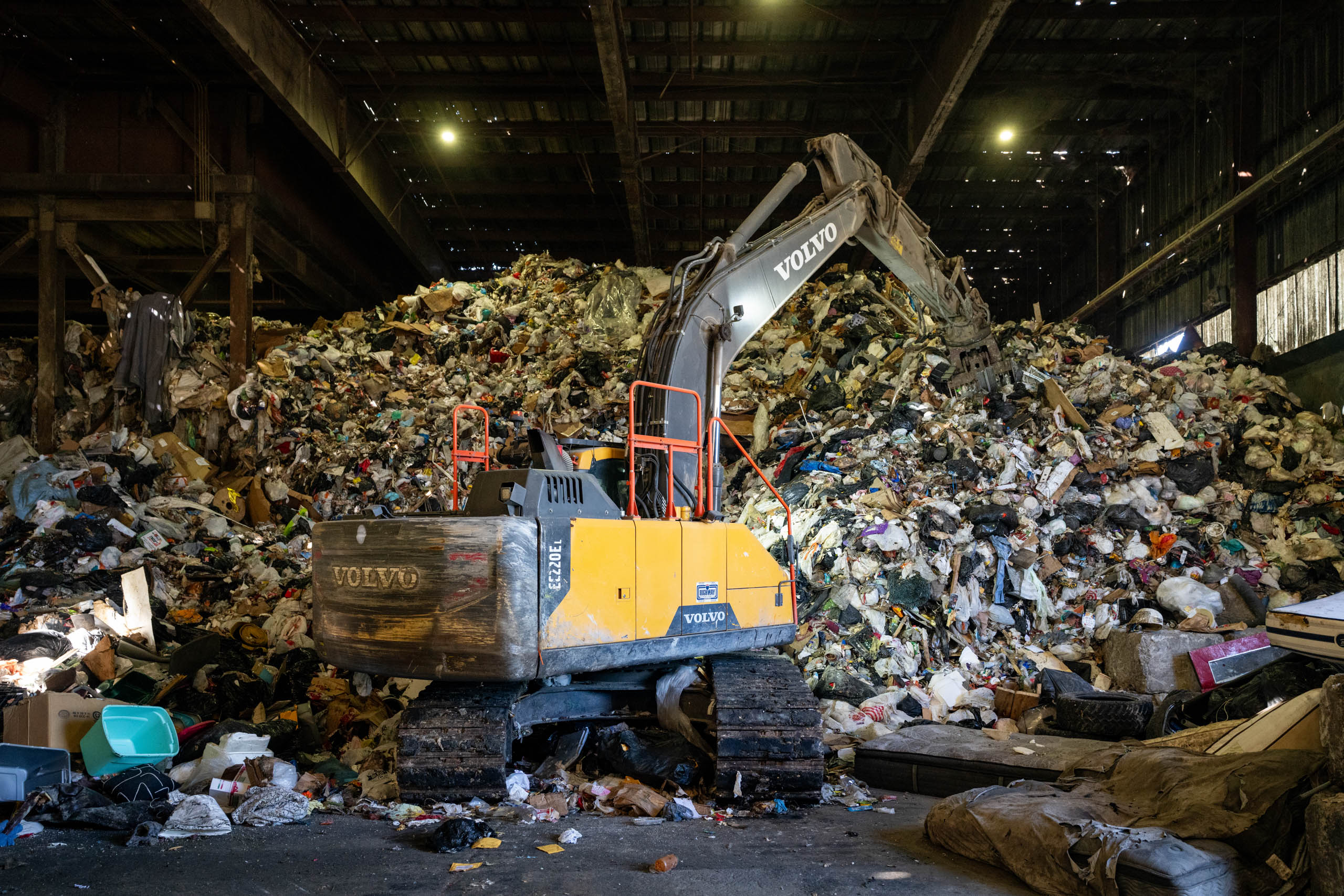 A yellow Volvo excavator sits inside a large lcswma warehouse, surrounded by towering piles of mixed garbage and debris under a high, partially lit roof.