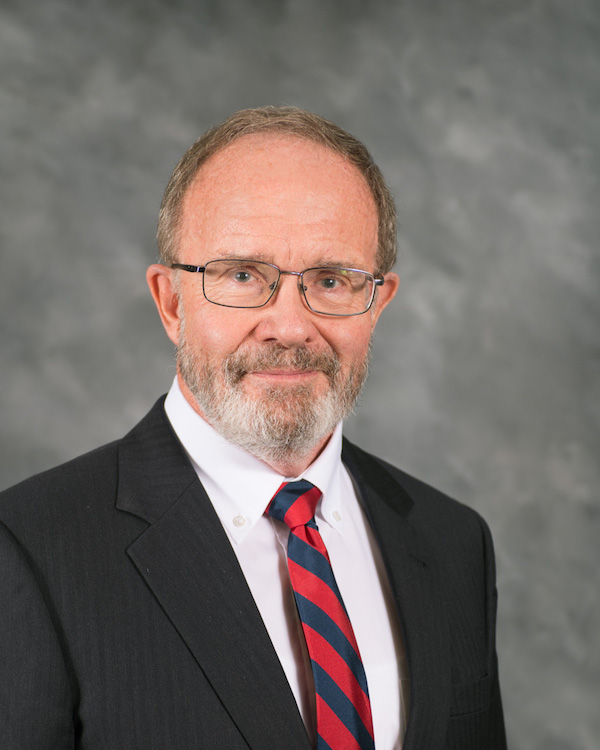A middle-aged man with short gray hair, glasses, and a trimmed beard, wearing a dark suit, white shirt, and red-and-blue striped tie, poses against a gray mottled background.