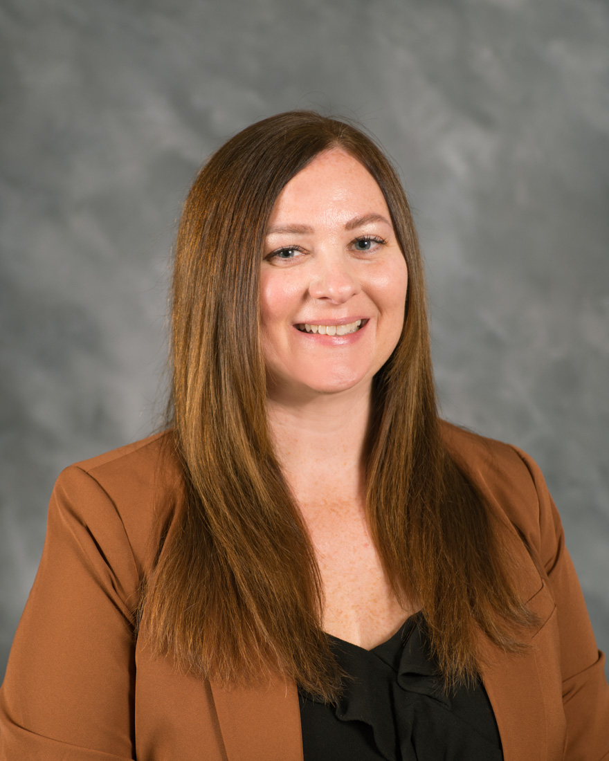 A woman with long straight brown hair wearing a brown blazer and black blouse smiles in front of a gray mottled background.