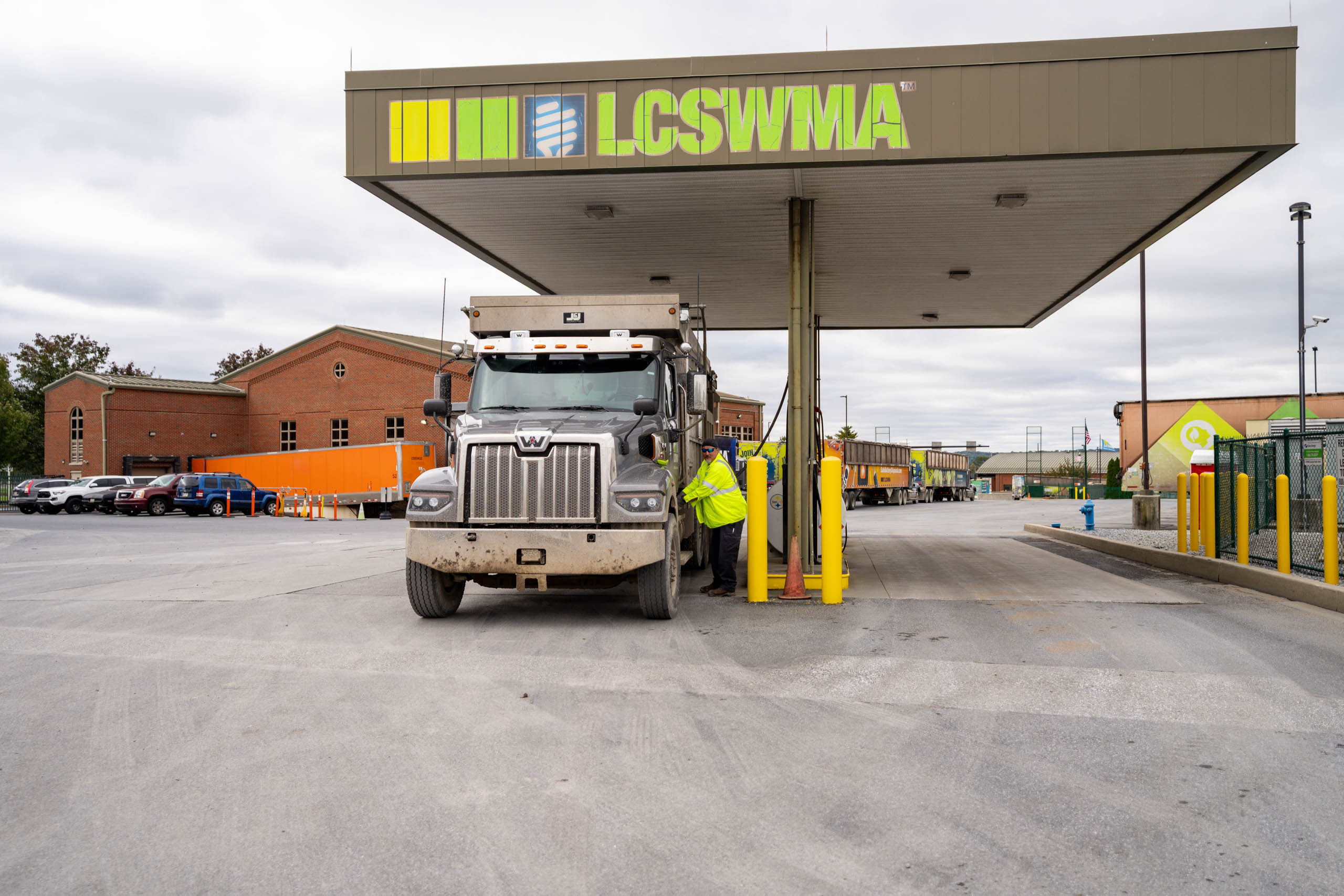 A person in a yellow safety jacket stands beside a large truck at an LCSWMA weigh station, highlighting the role of waste management within the waste ecosystem, with brick buildings and parked vehicles under a cloudy sky.