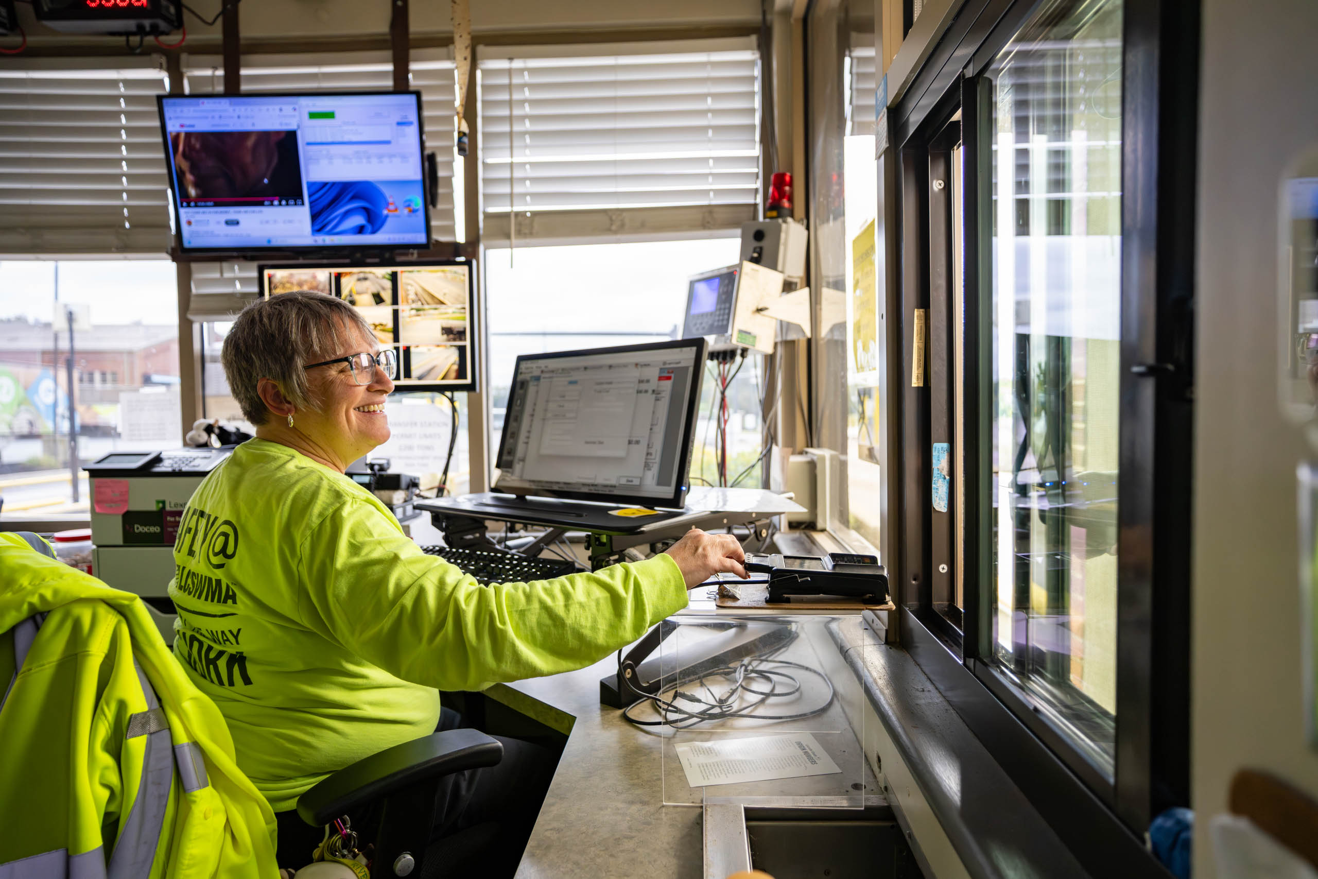 lcswma woman in a bright yellow safety shirt sits at a desk with computer monitors and paperwork, smiling as she manages renewable energy operations in a control room with large windows and multiple screens on the walls.