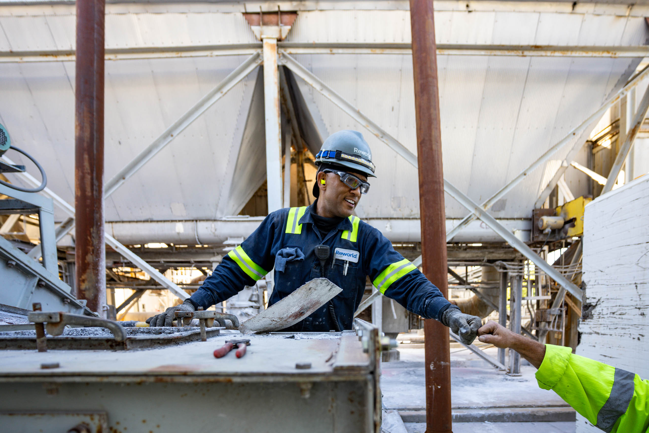 A lcswma worker in safety gear smiles while handing an object to a coworker, highlighting the importance of partnerships at an industrial worksite with metal beams and equipment in the background.