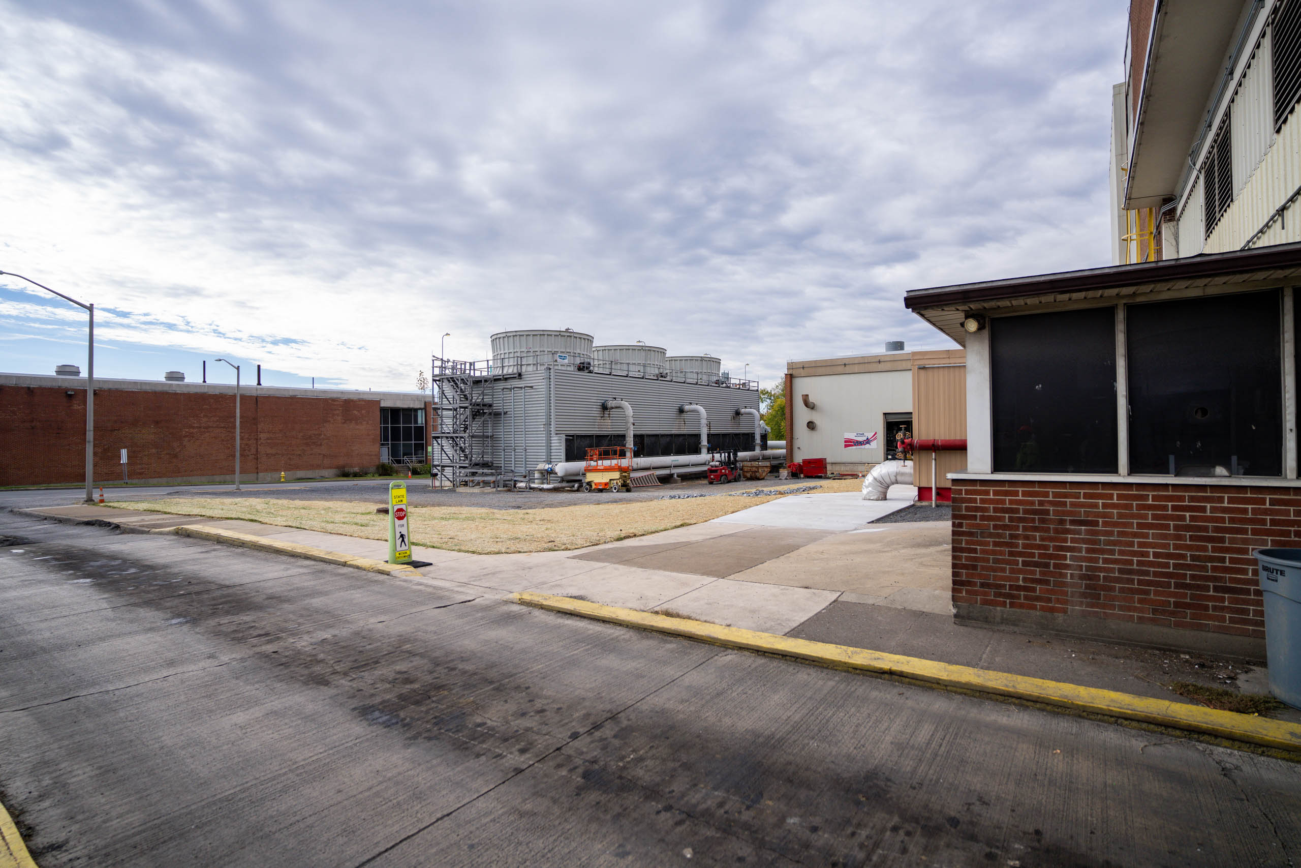 A paved industrial area with large gray cooling towers, scaffolding, and equipment under a cloudy sky. Brick buildings and a small booth are visible in the foreground and background, offering potential for additional resources on site.