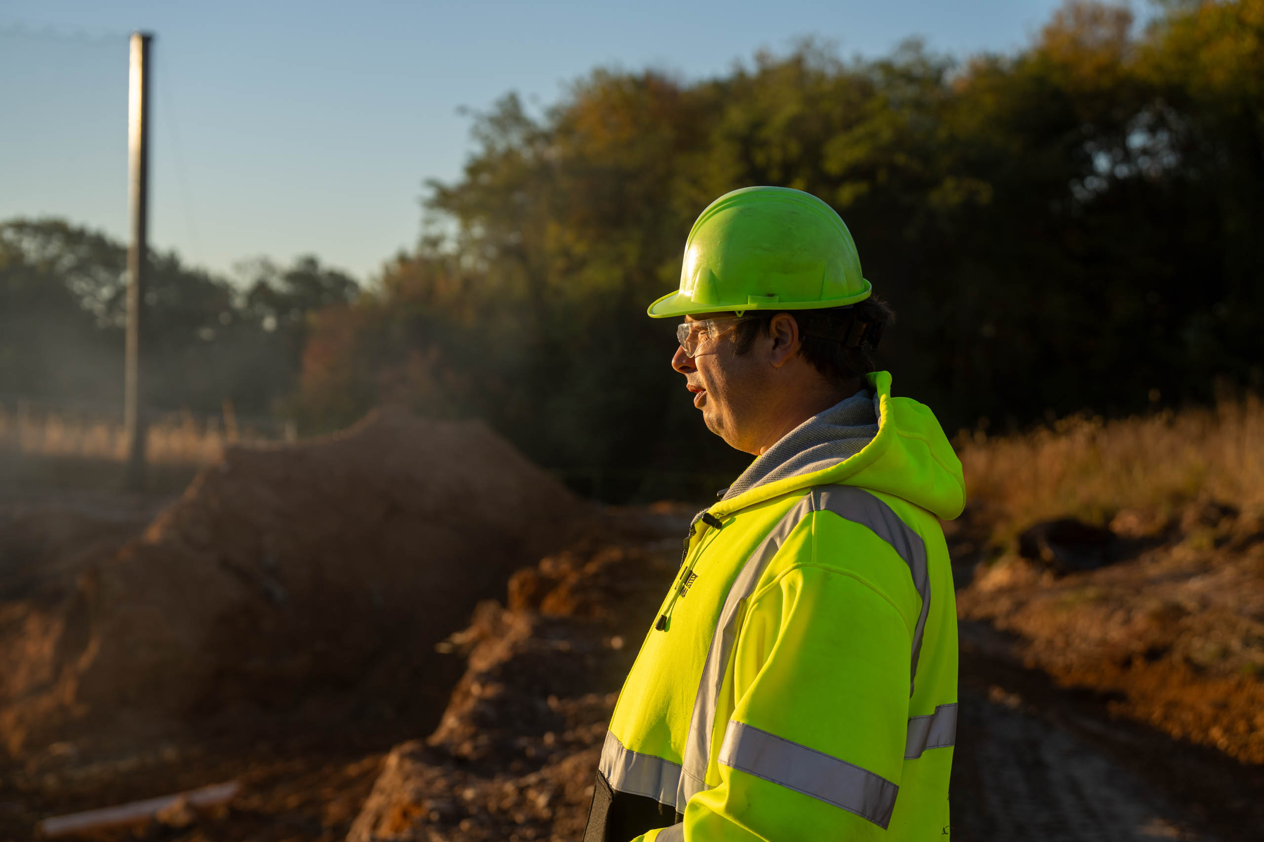 A lcswma worker wearing a neon yellow safety jacket and hard hat stands outdoors at a worksite, with dirt piles and trees in the background, in the early morning or late afternoon light.