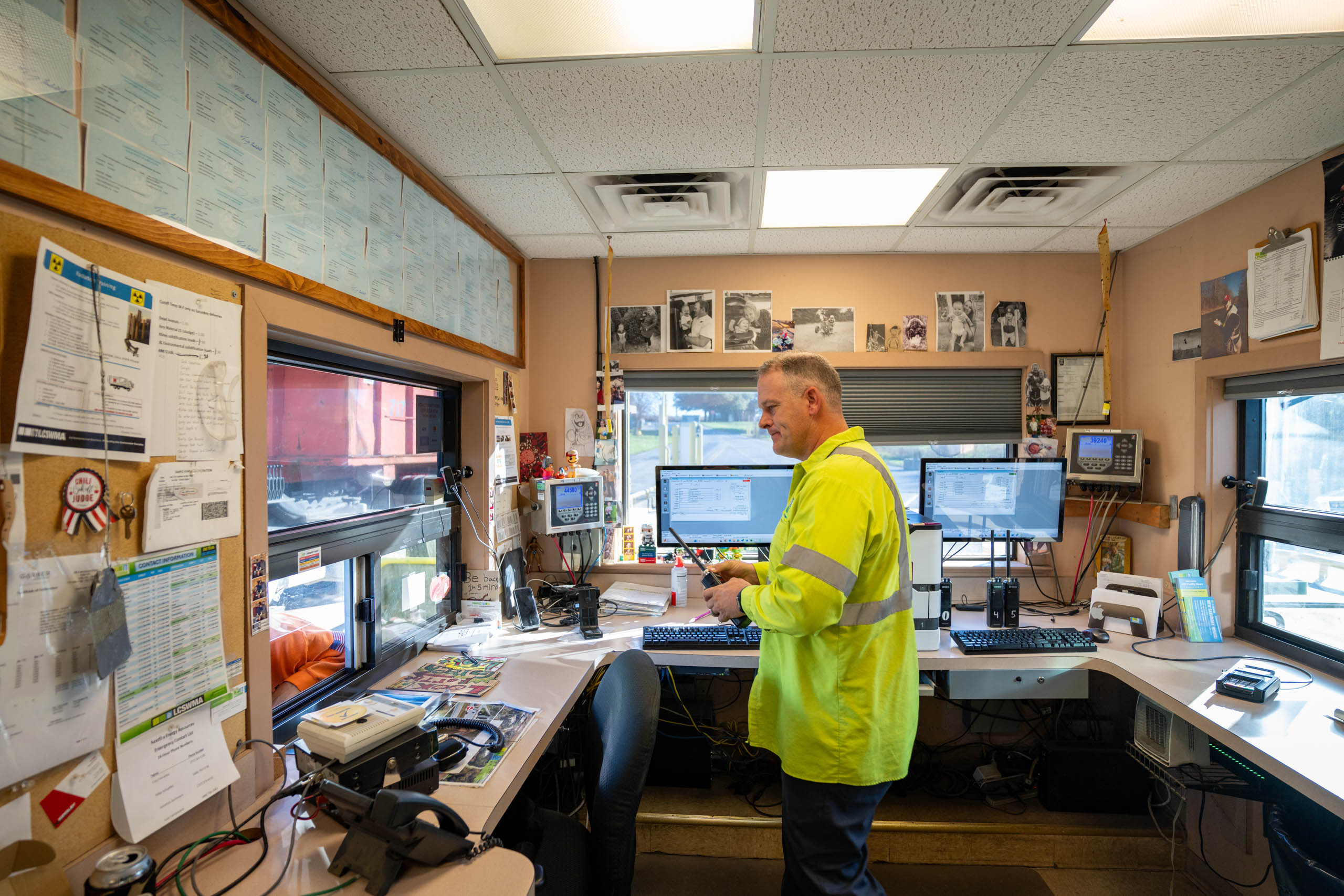 lcswma manager in a yellow high-visibility jacket stands in an office with multiple computer monitors, paperwork, photos on the walls, and access to additional resources as he appears to work or monitor information.