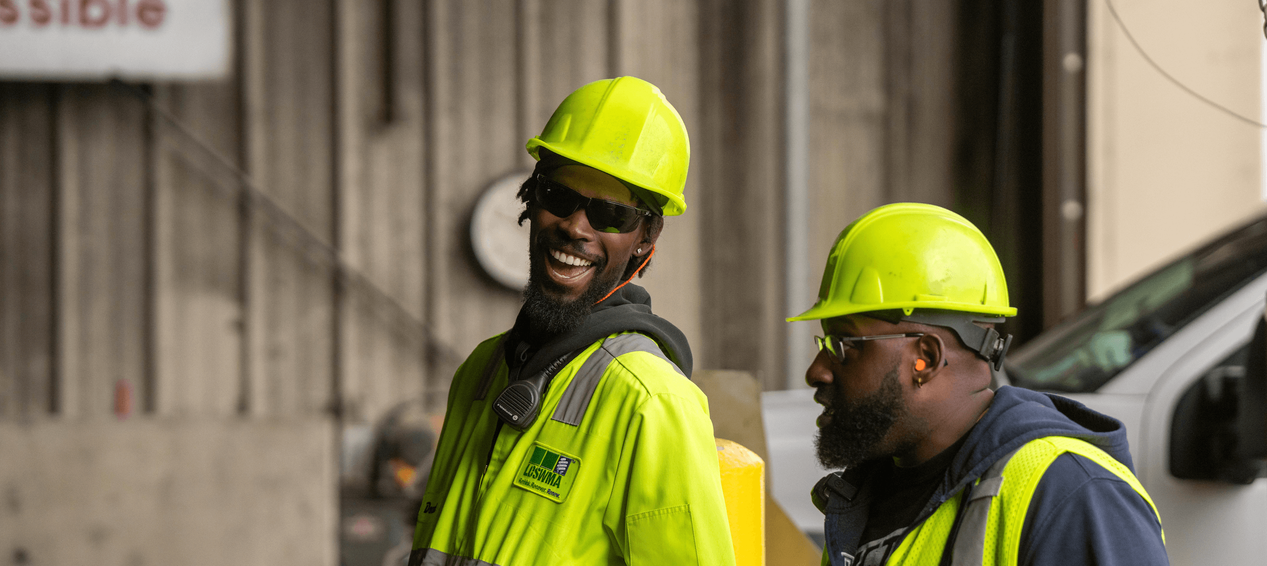 Two trash dump workers wearing neon yellow safety gear and hard hats stand together, one smiling broadly while the other looks ahead. Both wear sunglasses and are in an industrial setting.
