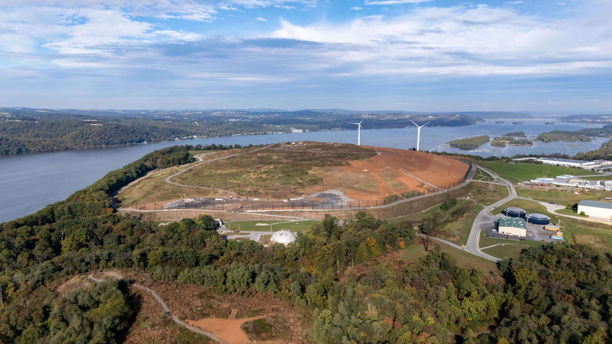 Aerial view of a large grassy hill with LCSWMA Facilities and construction areas, surrounded by water and forest. Two wind turbines stand on the hill, with buildings and a road in the foreground under a partly cloudy sky. Virtual Facility Tours available.