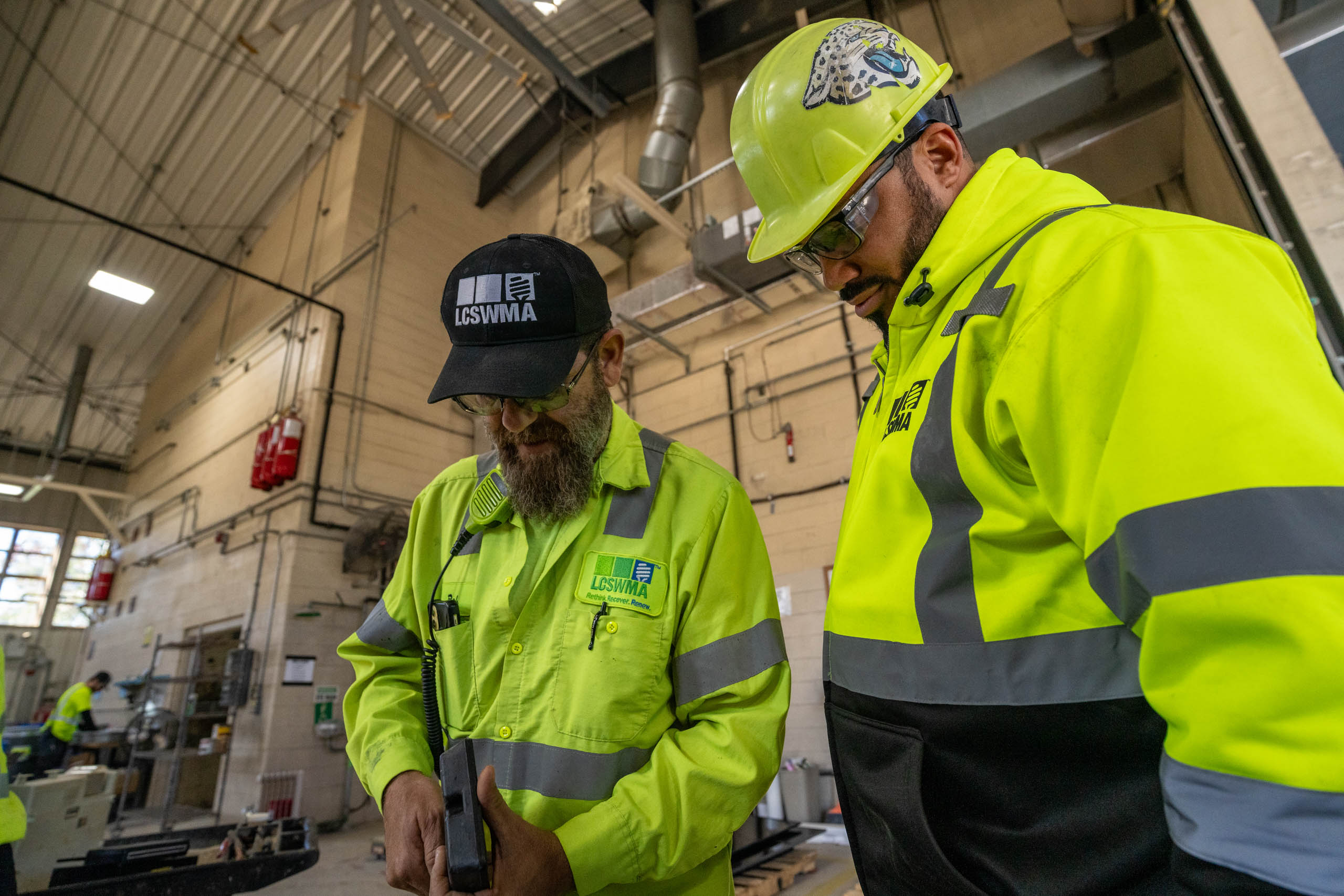 Two LCSWMA workers wearing high-visibility jackets and safety gear stand inside an industrial facility, focusing on a handheld device. The indoor setting has exposed pipes and industrial equipment in the background.