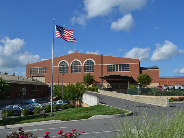 A large brick building with arched windows, an American flag flying in front, and several cars parked in a lot on a sunny day with blue sky and scattered clouds—explore more through Virtual Facility Tours of LCSWMA Facilities.