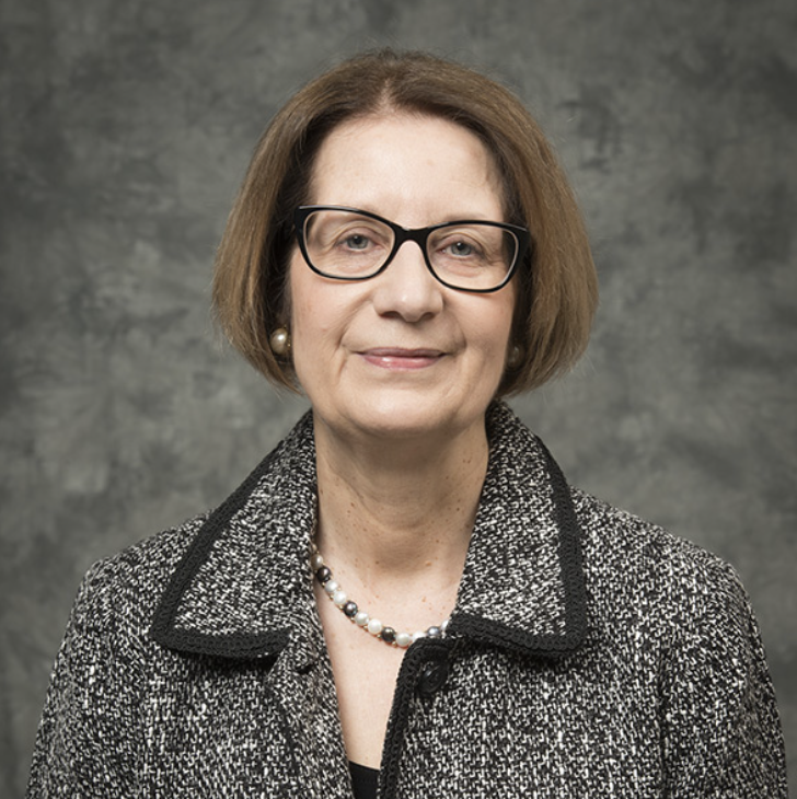 A middle-aged woman with short brown hair, wearing glasses, a gray textured jacket, a pearl necklace, and pearl earrings, stands in front of a gray mottled background, smiling slightly.
