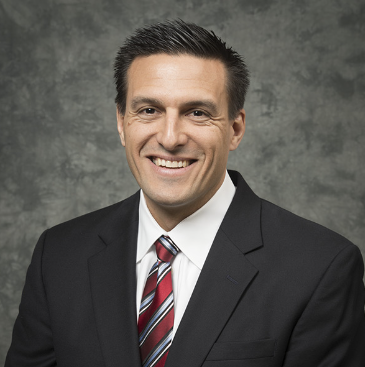 Steve Dzurik, dressed in a dark suit, white shirt, and red striped tie, smiles at the camera in front of a gray textured background.