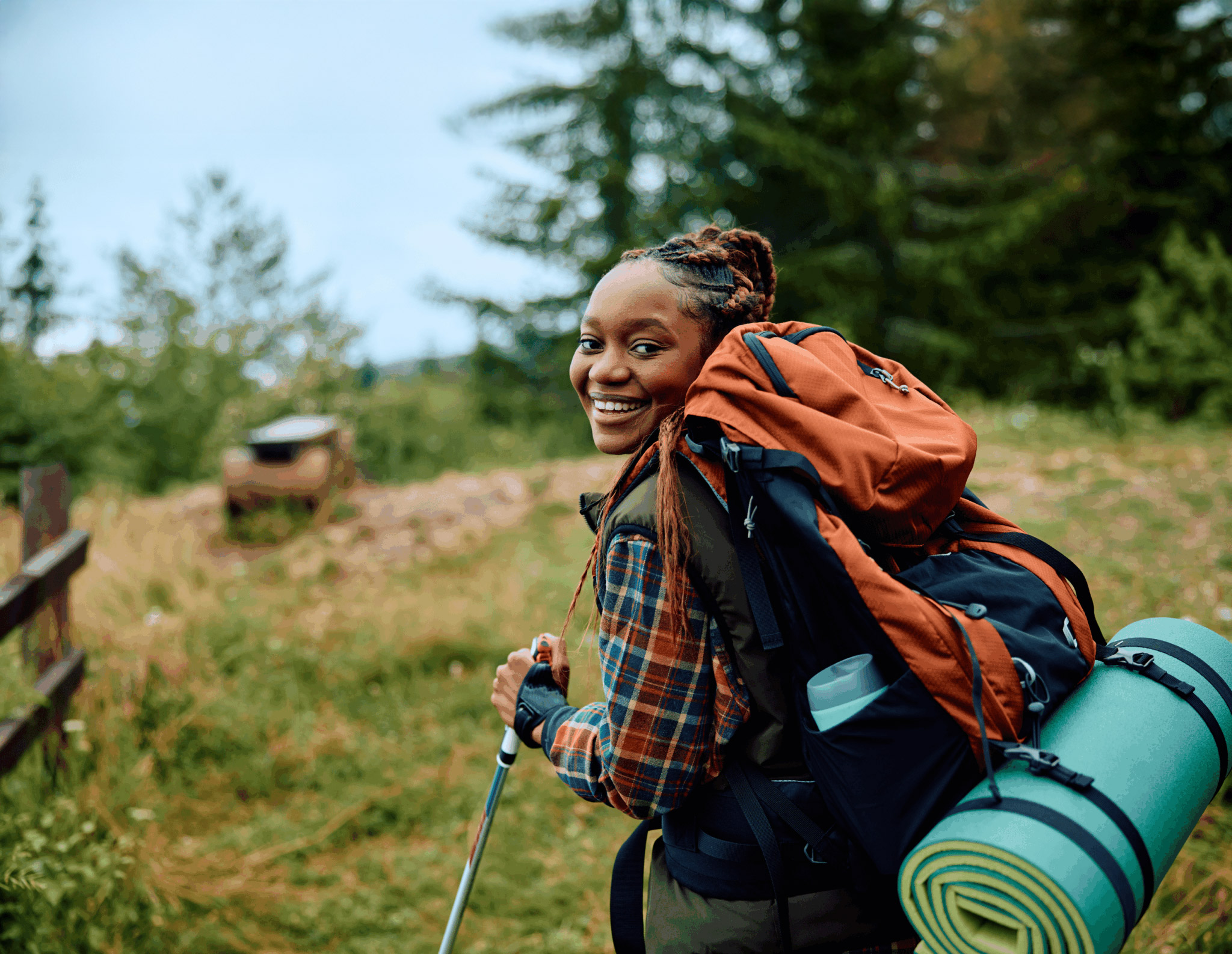 A woman with a large backpack and camping gear smiles at the camera while hiking on a forest trail, surrounded by greenery and trees.